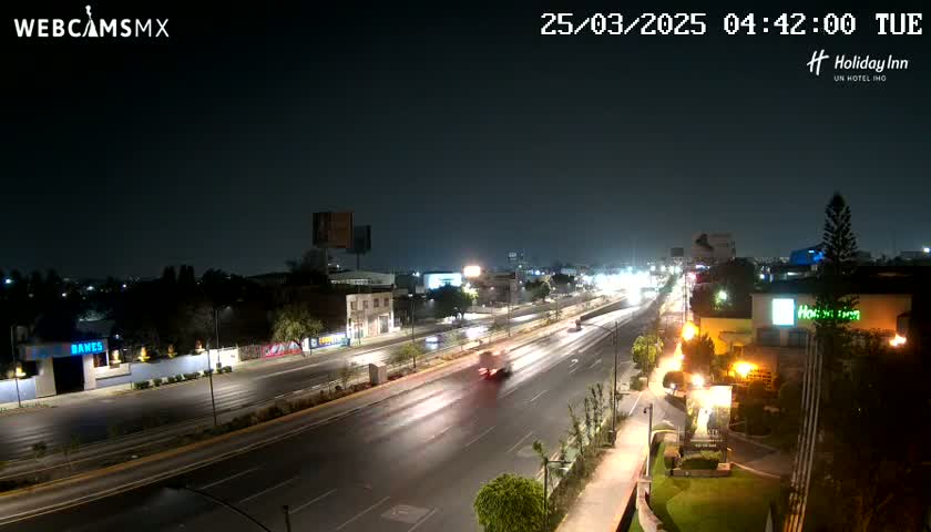 A nighttime view of a multi-lane highway with sparse traffic, surrounded by city buildings and landscaping.