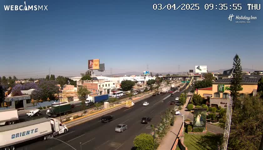 A sunny day reveals a multi-lane highway with moderate traffic, flanked by buildings, businesses, and landscaping.