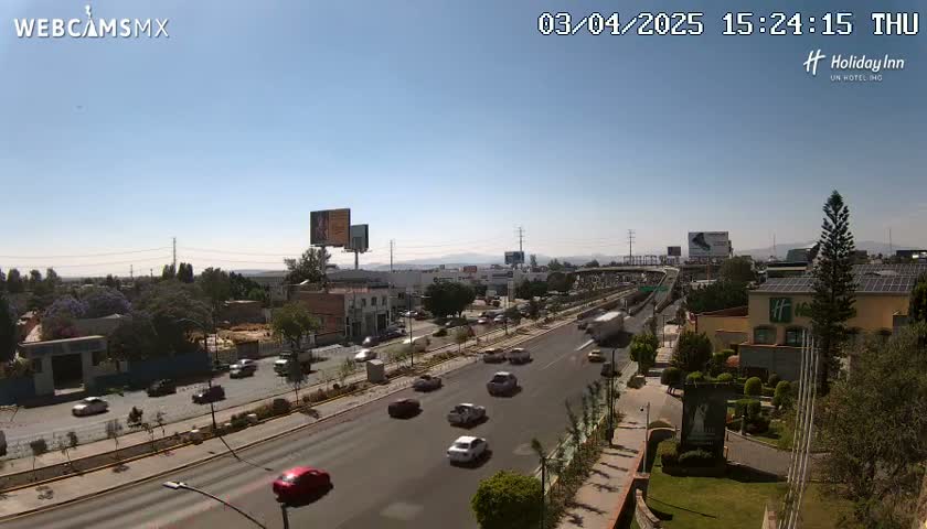 A sunny day shows a highway with moderate traffic, lined with buildings and landscaping, under a clear blue sky.