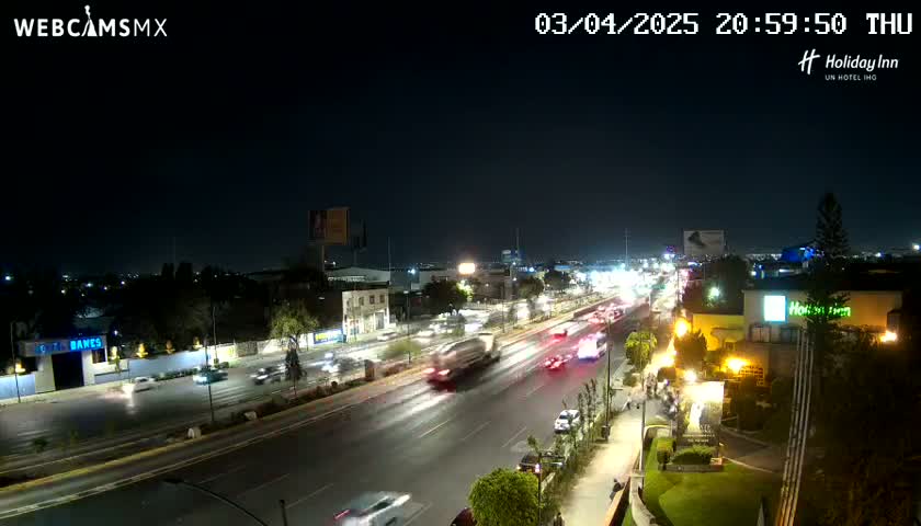 A nighttime view of a multi-lane road with moderate traffic, surrounded by buildings and illuminated by streetlights.