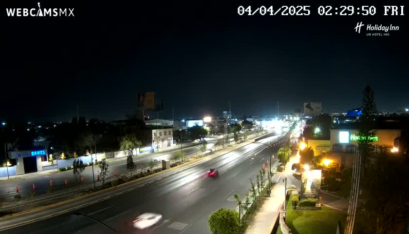 A nighttime view of a multi-lane highway with sparse traffic, surrounded by buildings and vegetation under a clear, dark sky.