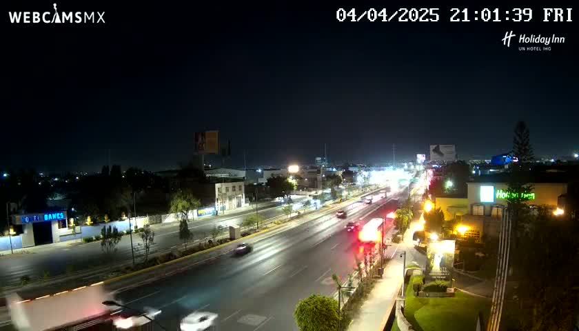 A nighttime view of a multi-lane road with cars driving in both directions, lined with buildings and trees under a clear, dark sky.