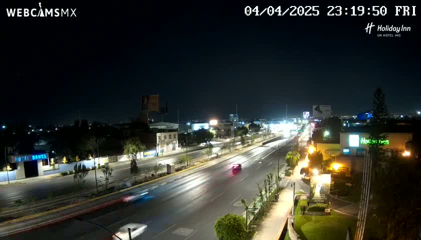 A nighttime, high-angle view of a multi-lane road with cars driving on it, surrounded by buildings and illuminated by streetlights.