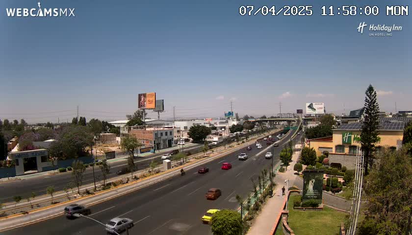 A multi-lane highway with moderate traffic flows through a city under a clear, sunny sky.