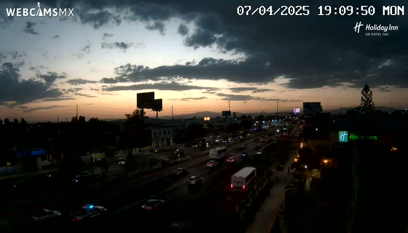 A partly cloudy sunset illuminates a multi-lane highway with moderate traffic, buildings, and billboards lining the roadway.