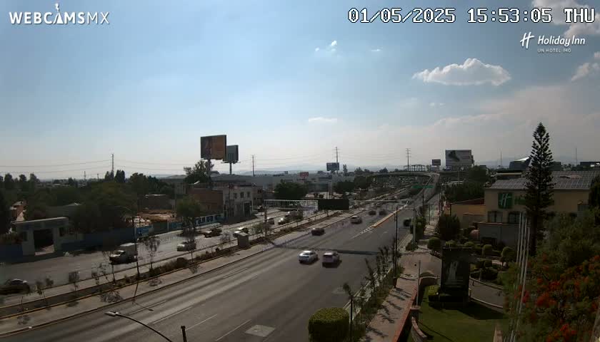 A partly sunny day reveals a multi-lane highway with sparse traffic, flanked by buildings and landscaping, extending toward a distant mountain range.