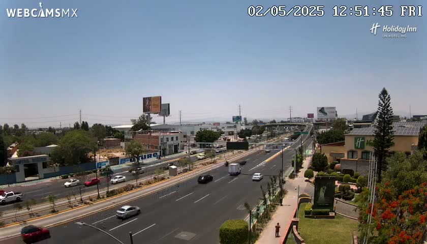 A sunny, clear day overlooks a multi-lane highway with moderate traffic, surrounded by buildings, landscaping, and billboards.