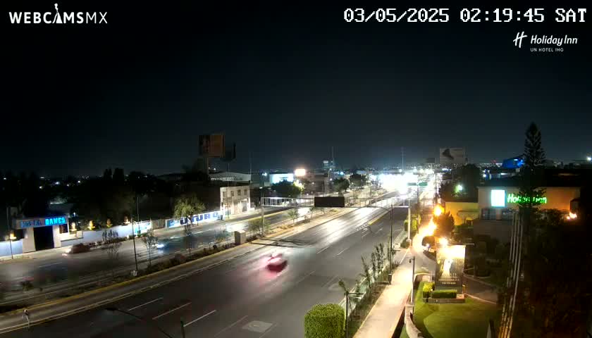 A nighttime view of a multi-lane road with sparse traffic, surrounded by buildings and greenery under a clear sky.