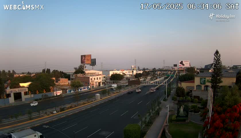 A highway with light traffic is seen at dawn under a clear, pale sky, with buildings and vegetation lining the roadside.