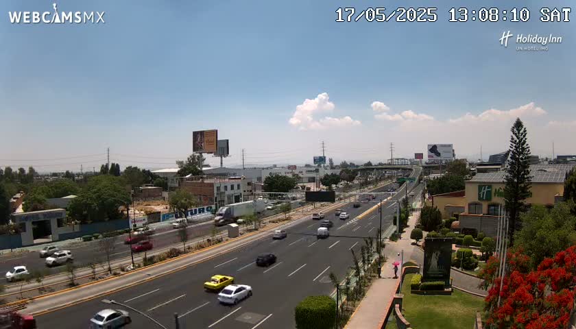 A partly cloudy day reveals a multi-lane highway with light traffic, surrounded by urban development including buildings and billboards.
