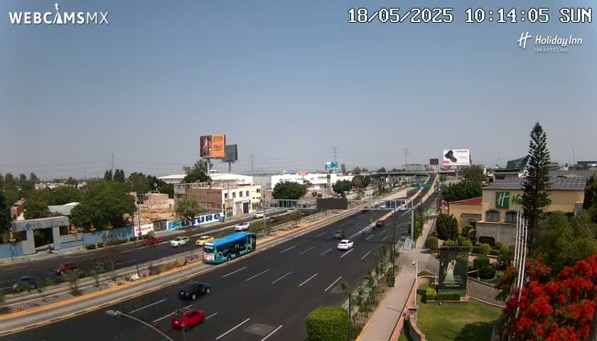 A sunny day shows a multi-lane highway with several vehicles, including a bus, surrounded by buildings, billboards, and lush green landscaping.