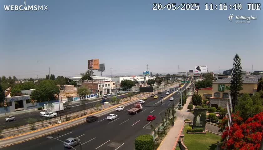 A highway scene on a sunny day shows numerous vehicles driving on a multi-lane road, with buildings, greenery, and billboards lining the sides.