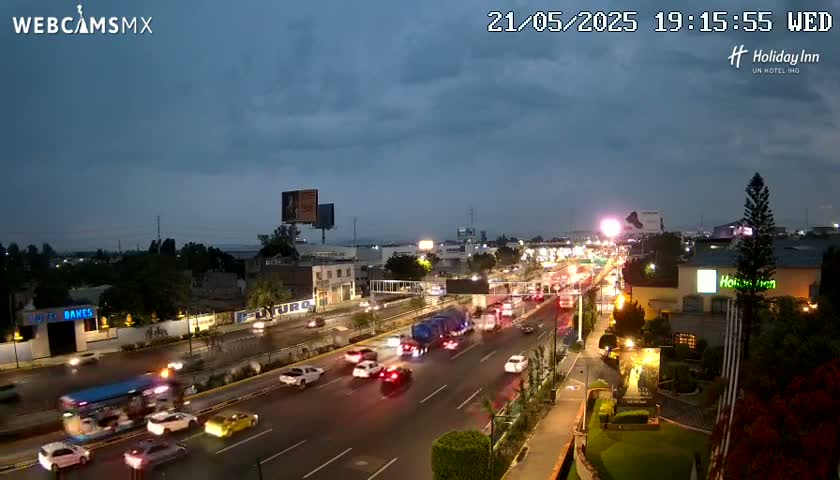 A nighttime, overcast view of a multi-lane highway with moderate traffic, surrounded by buildings and landscaping.