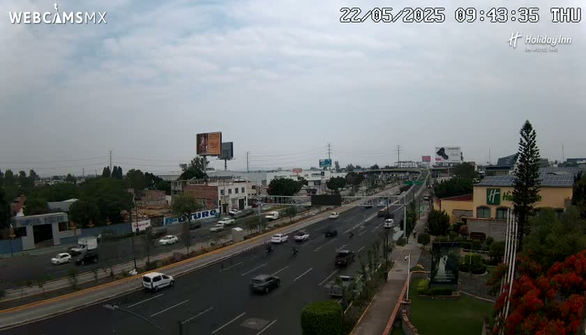 A multi-lane highway with moderate traffic is shown under an overcast sky, with buildings and greenery visible on either side.