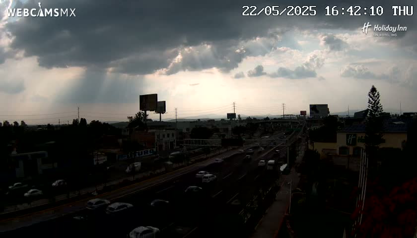 A highway with moderate traffic is seen under a partly cloudy sky with sunbeams breaking through the clouds.