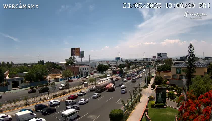A partly sunny day reveals a highway scene with moderate traffic, buildings, and lush green landscaping.