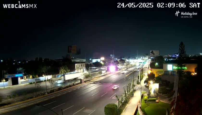 A nighttime view of a multi-lane highway with sparse traffic, surrounded by buildings and landscaping under clear skies.