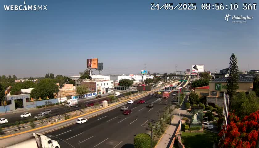 A highway with moderate traffic flows under a clear, sunny sky, surrounded by buildings and greenery.