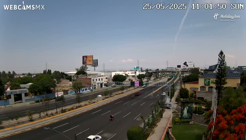 A mostly sunny, clear day overlooking a multi-lane highway with light traffic, surrounded by buildings, greenery, and billboards.