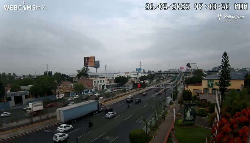 A highway with moderate traffic is shown under an overcast sky, with buildings and greenery visible on either side.