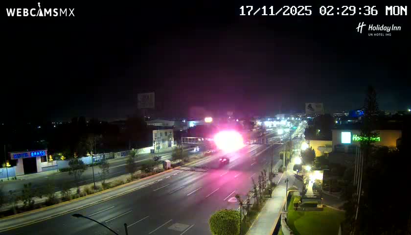 A clear night view shows a multi-lane city street illuminated by numerous streetlights and a prominent bright pink glow from a building or advertisement, with blurred vehicles moving along the road under a dark, clear sky.