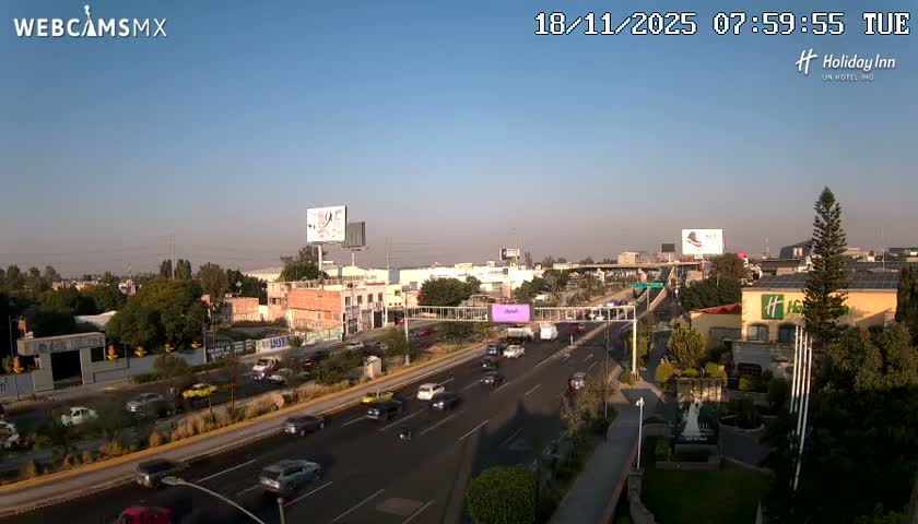 A clear night view shows a multi-lane city street illuminated by numerous streetlights and a prominent bright pink glow from a building or advertisement, with blurred vehicles moving along the road under a dark, clear sky.
