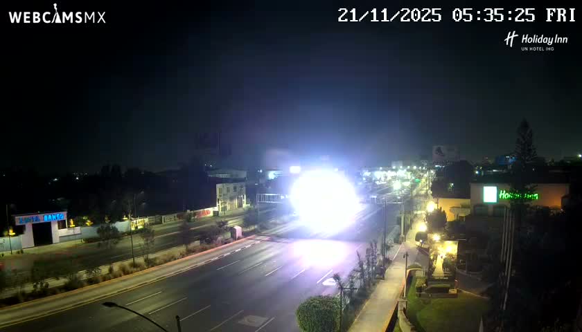 The image displays a clear night scene of a city road lined with buildings and illuminated by numerous streetlights, with a blinding bright light source dominating the middle distance.