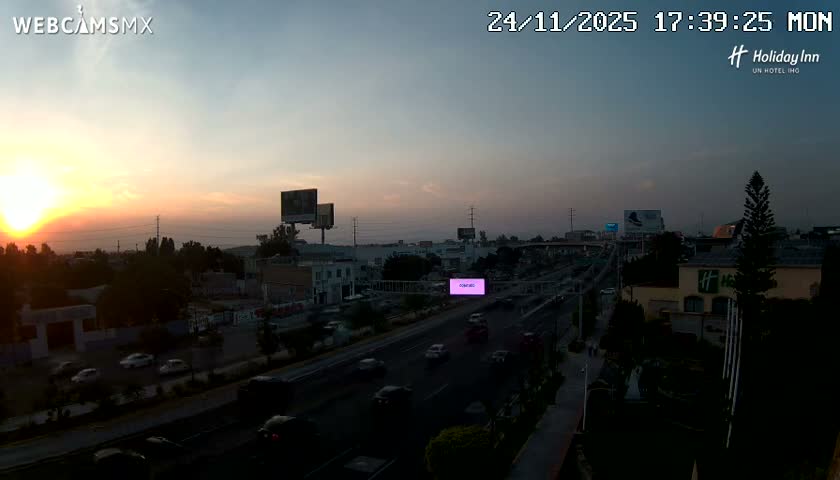 A clear sunset bathes an urban scene featuring a multi-lane road with active traffic, surrounded by buildings and numerous trees, under a sky transitioning from bright orange to dusky blue.