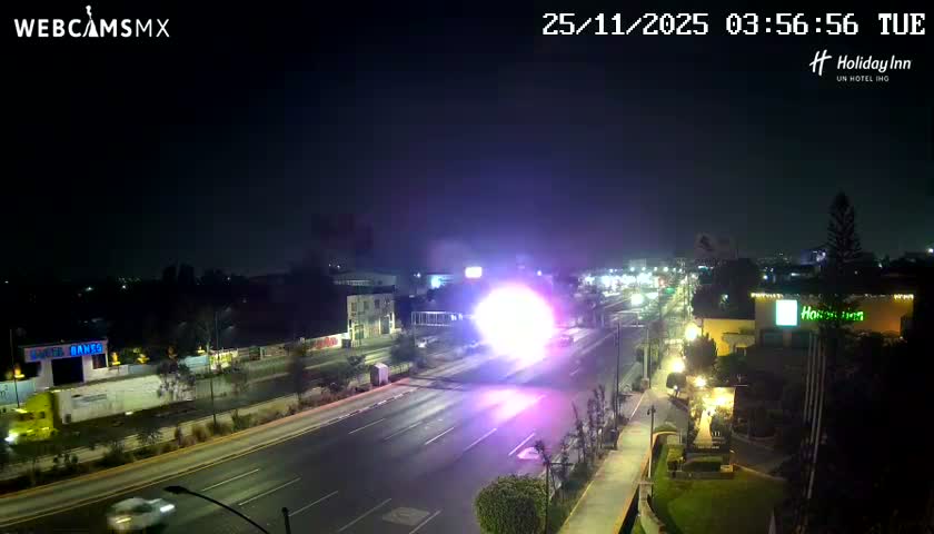 A wide road at night is brightly illuminated by a brilliant, spherical purple-white flash or explosion in the center, with blurred vehicle lights and various buildings visible under a clear, dark sky.