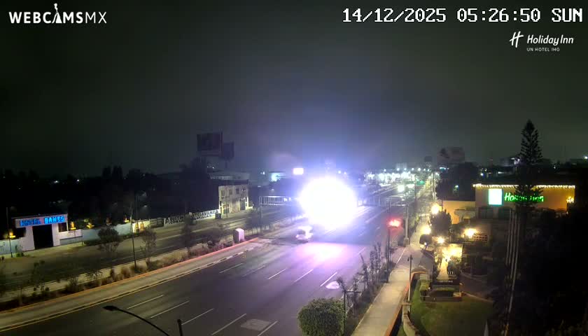 A wide road at night is brightly illuminated by a brilliant, spherical purple-white flash or explosion in the center, with blurred vehicle lights and various buildings visible under a clear, dark sky.