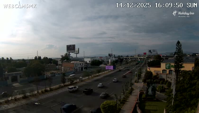 A wide road at night is brightly illuminated by a brilliant, spherical purple-white flash or explosion in the center, with blurred vehicle lights and various buildings visible under a clear, dark sky.