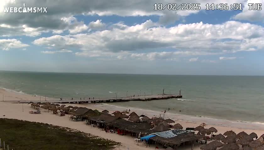 A partly cloudy day at a beach shows a long pier extending into a calm sea, with numerous beach umbrellas and thatched-roof structures lining the sandy shore.