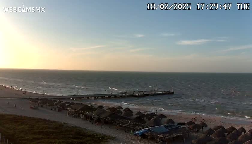 A beach with a pier extends into a moderately wavy ocean under a partly cloudy sunset sky.