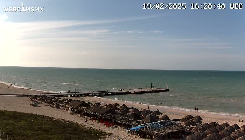 A beach with numerous thatched-roof structures and a pier extends into a calm, light-blue ocean under a mostly sunny sky.