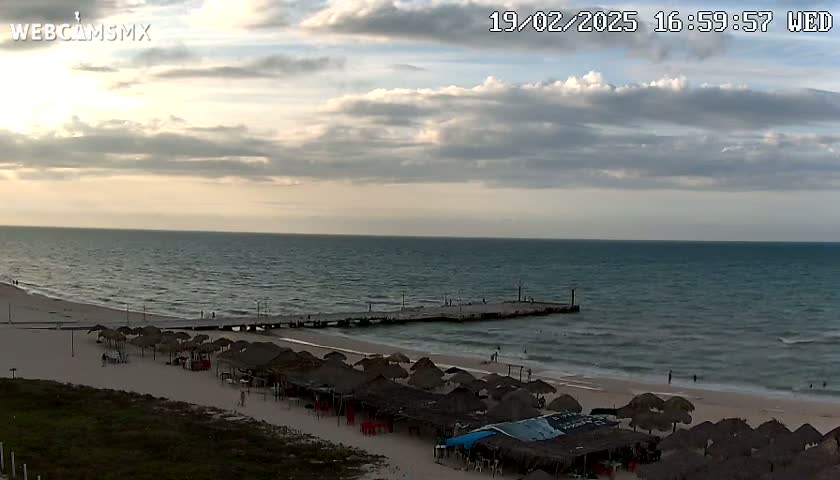 A partly cloudy sunset over a beach with calm waves, thatched-roof umbrellas, and a pier.
