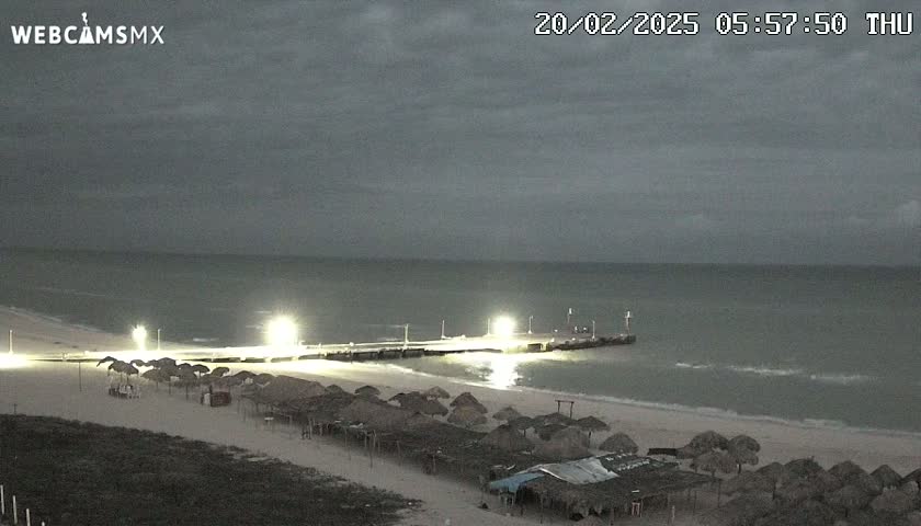 A beach at night under an overcast sky shows a pier lit with lights, beach umbrellas, and a calm sea.