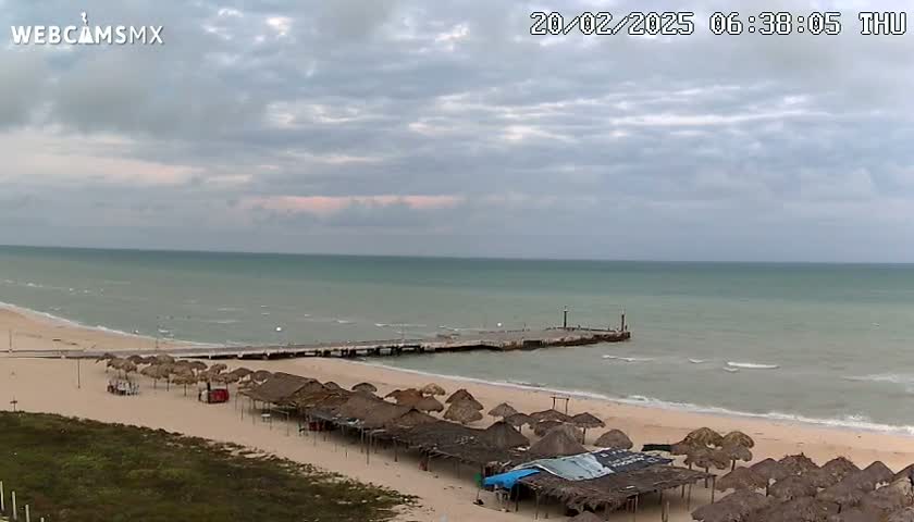 A beach with calm, light-green water under a mostly cloudy sky features a pier and numerous thatched-roof structures on the sand.