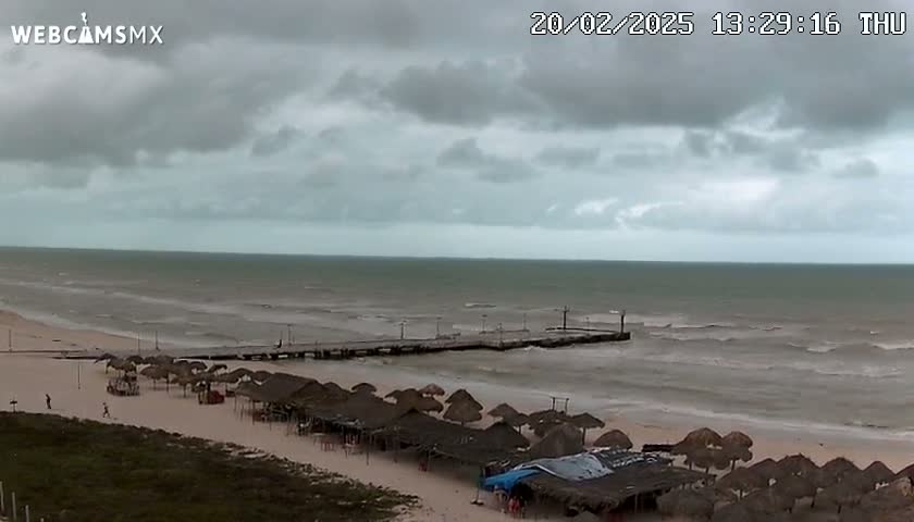 A beach with numerous thatched umbrellas under an overcast sky and a somewhat choppy sea with a pier extending into the water.