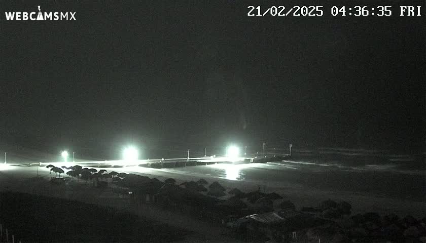A nighttime beach scene shows a pier lit by several bright lights, with calm waves lapping the shore and rows of closed beach umbrellas in the foreground.
