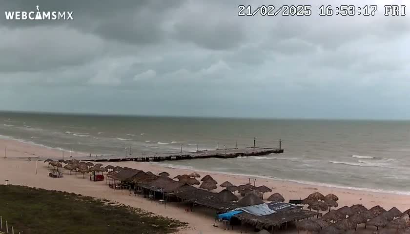 A beach with numerous thatched umbrellas and a pier extends into a somewhat choppy sea under a cloudy sky.