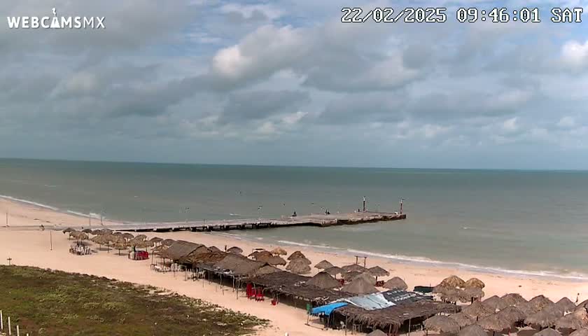 A partly cloudy day shows a beach with numerous thatched umbrellas and a long pier extending into a calm sea.