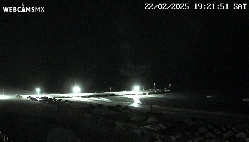 A nighttime beach scene shows a pier lit by several lights, with dark structures on the sand in front of it.