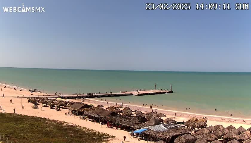A sandy beach with thatched-roof structures and a pier extends into a calm, light-green ocean under a clear blue sky.