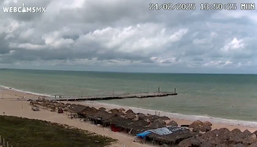 A beach with numerous thatched-roof structures is visible under a cloudy sky, with a pier extending into a calm, light-green ocean.