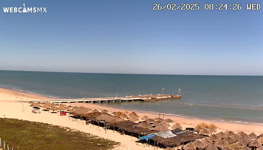 A long pier extends into a calm, blue ocean from a sandy beach lined with numerous thatched-roof structures.