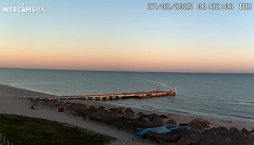 A long pier extends into a calm ocean under a pale pink and orange sunset, with a sandy beach lined with thatched umbrellas below.