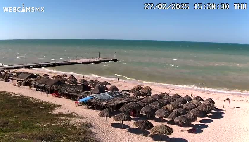 A sandy beach with numerous thatched umbrellas, a pier extending into a somewhat choppy sea under a clear blue sky.