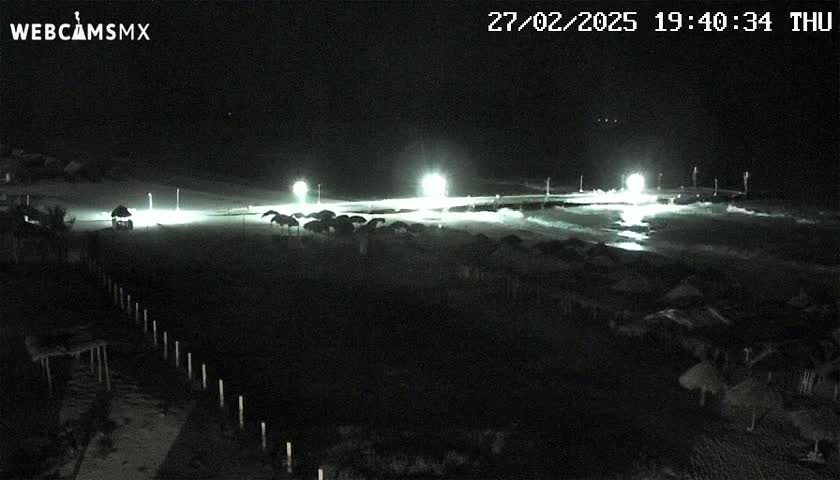 A nighttime beach scene shows a dark sandy area with several lights illuminating structures, likely umbrellas or beach shelters, under a clear, dark sky.