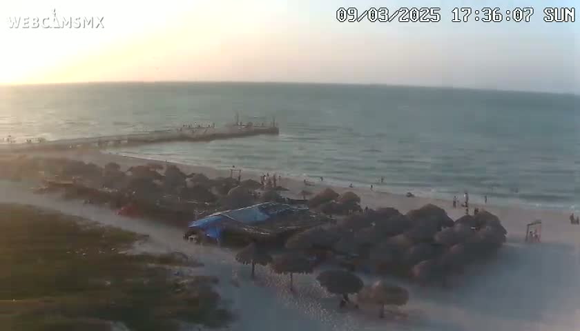 A beach scene at sunset shows a pier, calm ocean waves, a sandy beach with numerous thatched umbrellas, and a few people scattered about.