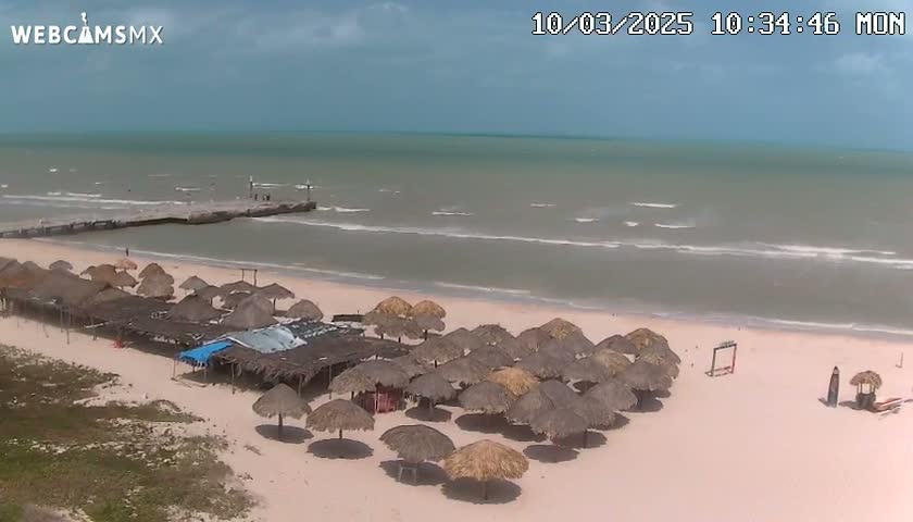 A sandy beach with numerous thatched umbrellas, a pier extending into a somewhat choppy sea under a partly cloudy sky.
